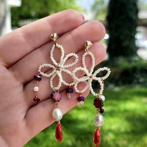 Può includere: Un paio di orecchini pendenti con motivi floreali in perline bianche, perni a forma di cuore dorati e pendenti che includono perle d'acqua dolce bianche, perline di vetro rosso scuro e ciondoli rossi a forma di goccia su filo dorato.