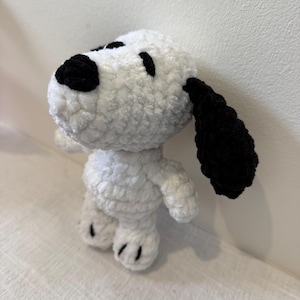 Peut inclure: Une figurine Snoopy au crochet faite à la main, avec un corps blanc et des détails noirs sur les oreilles, le nez et les yeux. Le jouet en peluche est debout, avec de petits bras et pieds. La laine douce et texturée donne à la figurine un aspect câlin.