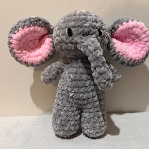 May include: A plush elephant toy crafted from soft, grey velvet-textured yarn. The elephant features large, rounded ears with bright pink inner detailing, a trunk, and two small black eyes. The body has a thick, crocheted appearance throughout.