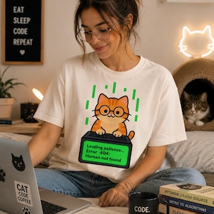 Op de afbeelding: Crèmekleurig T-shirt met een cartoonkat met een bril en een computerfoutmelding. De tekst op het shirt luidt: "Loading patience... Error 404: Human not found." Een zwarte mok met "CODE. COFFEE. REPEAT." is ook zichtbaar.