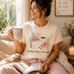 May include: A cream-colored t-shirt with the text "Booked, Busy & Blessed" and a graphic of a calendar, coffee cup, and book. The person is holding a mug that says "His Still is enough". The person is also holding a book.
