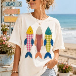 May include: An off-white short-sleeve t-shirt featuring a graphic print of three stylised fish. The fish are designed with fabric-like textures, including pink gingham, orange stripes, and yellow gingham patterns, all with blue fins and tails.
