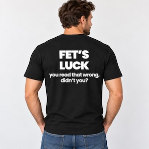 May include: A black short-sleeved t-shirt featuring white text on the back. The text reads FET'S LUCK followed by the sentence you read that wrong, didn't you? The shirt is worn with blue denim jeans, displaying a casual and simple design.