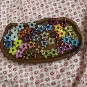 Könnte beinhalten: Ein Holztablett enthält eine Auswahl an glitzernden sternförmigen Verzierungen. Die Sterne sind in verschiedenen Farben gehalten, darunter Blau, Rosa, Gelb, Orange und Grün. Der Glitzer verleiht den Sternformen Glanz. Das Tablett liegt auf einem Stoff mit Blumenmuster.