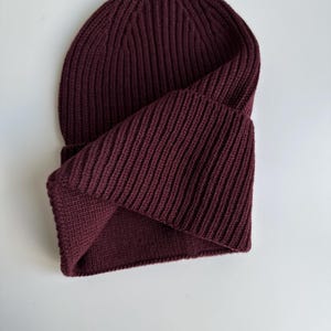 May include: A burgundy ribbed knit beanie hat. The hat is folded over, revealing the interior. The knit pattern runs vertically. The hat is made of a soft, warm material, perfect for cold weather.