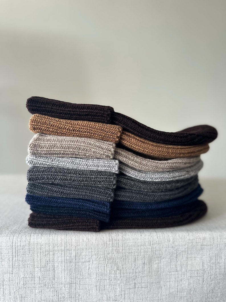 May include: A stack of various colored socks, including shades of brown, gray, and navy blue. The socks are neatly folded and stacked on top of each other, showcasing their different hues and textures. The socks appear to be made of a soft, knitted material.