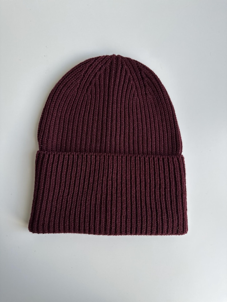 May include: A burgundy ribbed knit beanie hat. The hat is made of a soft, textured material and features a folded brim. The hat is a solid color and is suitable for cold weather.