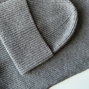 May include: Close-up of a gray knit beanie and matching scarf. The ribbed knit fabric is a solid gray color. The beanie is folded over, and the scarf is draped. The texture of the knitwear is visible.