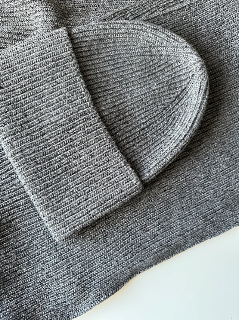 May include: Close-up of a gray knit beanie and matching scarf. The ribbed knit fabric is a solid gray color. The beanie is folded over, and the scarf is draped. The texture of the knitwear is visible.