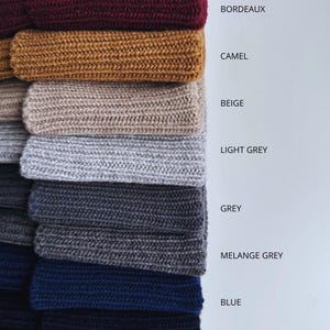 May include: A stack of knitted fabric samples in various colors, including pink, burgundy, camel, beige, light grey, grey, melange grey, blue, navy, and brown. Each sample is labeled with its corresponding color name.