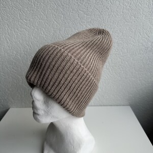 May include: A tan ribbed knit beanie hat displayed on a white mannequin head. The hat is made of a soft, textured material and has a folded brim. The beanie is a neutral color, suitable for various outfits.