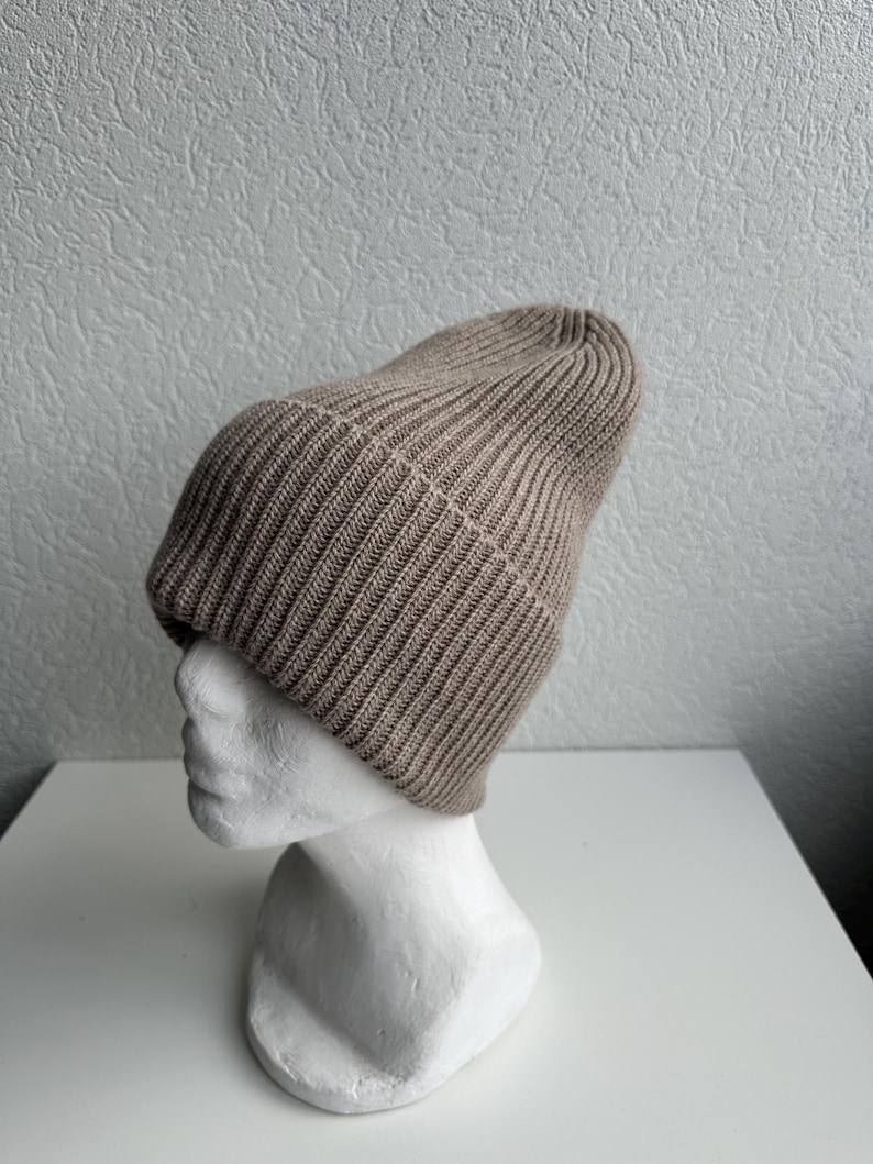 May include: A tan ribbed knit beanie hat displayed on a white mannequin head. The hat is made of a soft, textured material and has a folded brim. The beanie is a neutral color, suitable for various outfits.