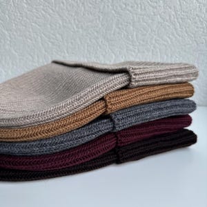 May include: A stack of six knitted beanies in various colors, including beige, brown, gray, and burgundy. The beanies are folded and stacked neatly, showcasing their ribbed texture and simple design. The hats are made of a soft, warm material.