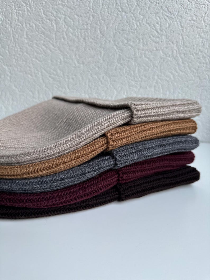 May include: A stack of six knitted beanies in various colors, including beige, brown, gray, and burgundy. The beanies are folded and stacked neatly, showcasing their ribbed texture and simple design. The hats are made of a soft, warm material.