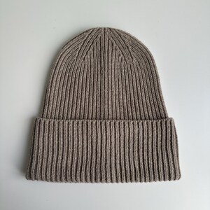 May include: A ribbed knit beanie in a neutral taupe color. The hat features a folded brim and a vertical ribbing pattern throughout. The beanie is made of a soft, warm material, suitable for cold weather.