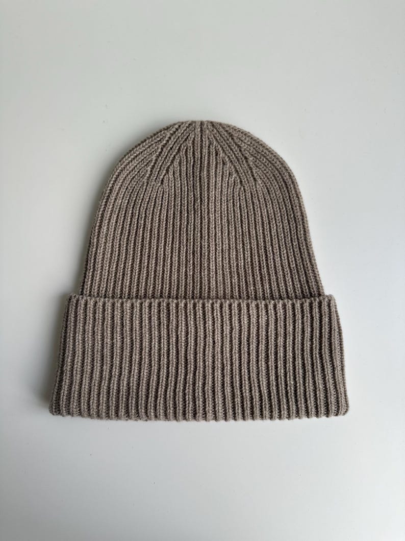 May include: A ribbed knit beanie in a neutral taupe color. The hat features a folded brim and a vertical ribbing pattern throughout. The beanie is made of a soft, warm material, suitable for cold weather.