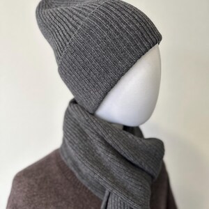 May include: A gray ribbed knit beanie and scarf set. The beanie is folded at the brim and the scarf is draped around a mannequin's neck. The set is made of a soft, textured material and is a neutral color.