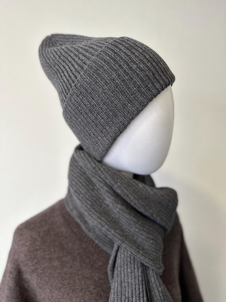 May include: A gray ribbed knit beanie and scarf set. The beanie is folded at the brim and the scarf is draped around a mannequin's neck. The set is made of a soft, textured material and is a neutral color.