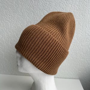 May include: A brown ribbed knit beanie hat. The hat is displayed on a white mannequin head. The beanie has a folded brim and a close-fitting design. The hat is made of a soft, warm material.