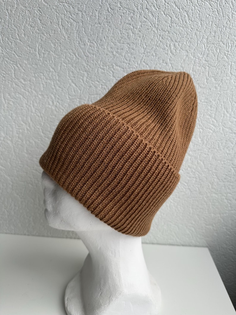 May include: A brown ribbed knit beanie hat. The hat is displayed on a white mannequin head. The beanie has a folded brim and a close-fitting design. The hat is made of a soft, warm material.
