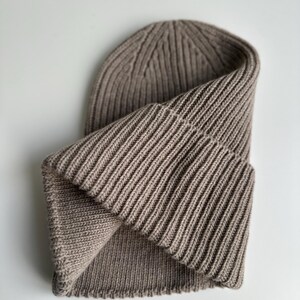 May include: A folded, ribbed knit beanie in a neutral taupe color. The hat is made of a soft, textured material and features a folded brim. The ribbed design runs vertically, creating a classic, versatile look. This beanie is suitable for cold weather.