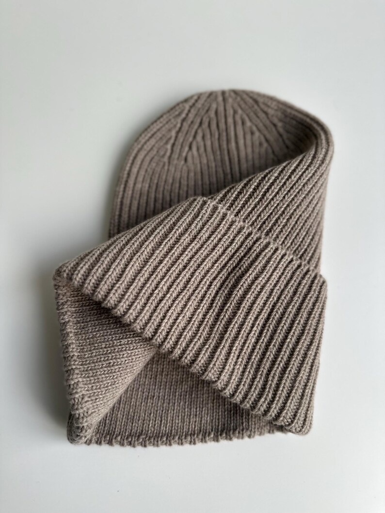 May include: A folded, ribbed knit beanie in a neutral taupe color. The hat is made of a soft, textured material and features a folded brim. The ribbed design runs vertically, creating a classic, versatile look. This beanie is suitable for cold weather.
