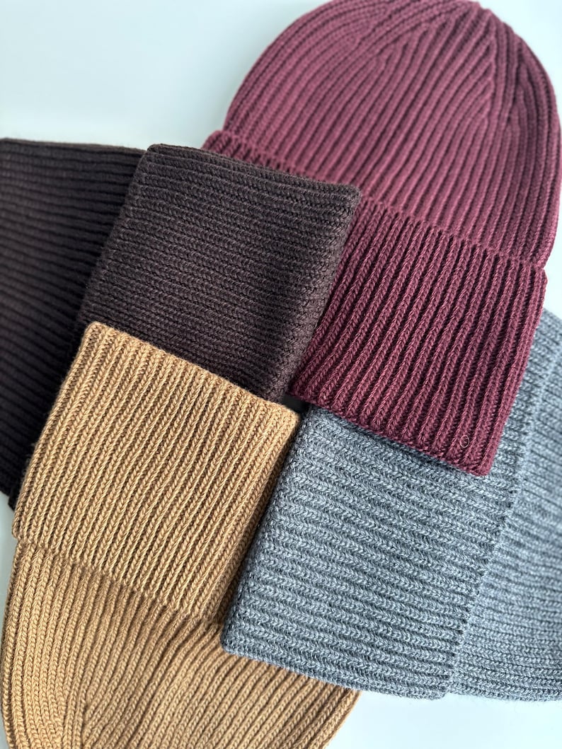 May include: A collection of ribbed knit beanies and matching scarves in various colors. The colors include a deep burgundy, dark brown, tan, and gray. The beanies are folded over at the brim.