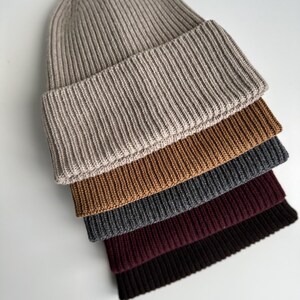 May include: A stack of ribbed knit beanies in various colors: beige, brown, gray, burgundy, and black. The hats are folded at the brim and arranged diagonally, showcasing the different shades and textures of the knitwear.