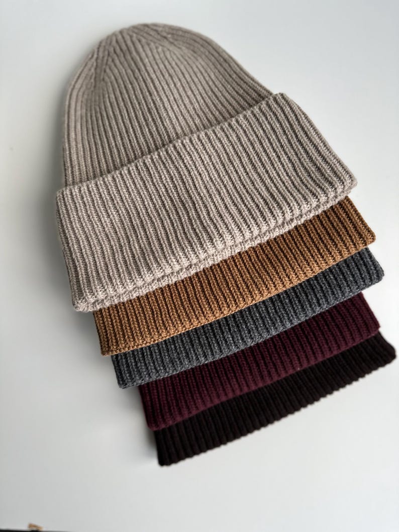 May include: A stack of ribbed knit beanies in various colors: beige, brown, gray, burgundy, and black. The hats are folded at the brim and arranged diagonally, showcasing the different shades and textures of the knitwear.