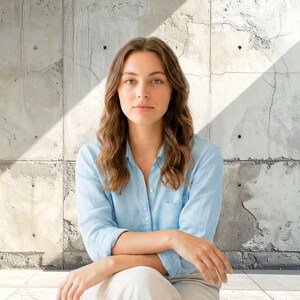 Puede incluir: Una mujer con una camisa azul claro abotonada y pantalones blancos está sentada frente a una pared de hormigón. La camisa tiene mangas largas y cuello. La mujer tiene el pelo largo, ondulado y castaño y mira directamente a la cámara.