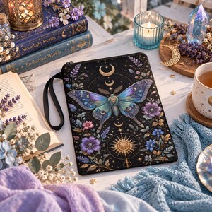Puede incluir: Un estuche rectangular negro con cremallera que presenta una ilustración central de una polilla con alas azules y moradas, rodeada de motivos florales, un sol dorado y símbolos de media luna. Incluye una correa de muñeca negra.