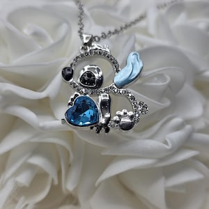 May include: A silver-toned pendant necklace featuring a cartoon character design. The pendant has a blue heart-shaped gem, black accents, and a light blue ear. The design is embellished with clear crystals.
