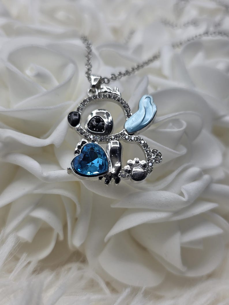 May include: A silver-toned pendant necklace featuring a cartoon character design. The pendant has a blue heart-shaped gem, black accents, and a light blue ear. The design is embellished with clear crystals.