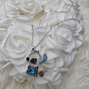 May include: Silver-toned necklace featuring a pendant shaped like a cartoon character. The pendant has a blue heart-shaped gem, blue accents, and is encrusted with clear crystals. The necklace is displayed on a bed of white roses.