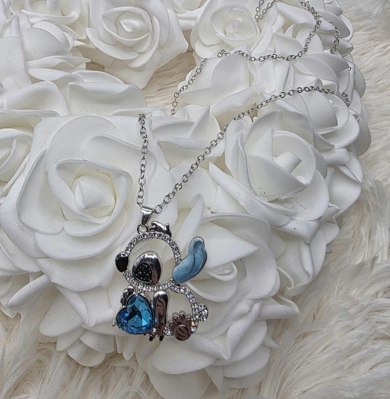 May include: Silver-toned necklace featuring a pendant shaped like a cartoon character. The pendant has a blue heart-shaped gem, blue accents, and is encrusted with clear crystals. The necklace is displayed on a bed of white roses.