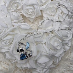 May include: A silver-toned necklace featuring a pendant of a cartoon character with a blue heart-shaped gem. The pendant is surrounded by clear crystals and rests on a bed of white roses.