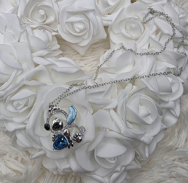 May include: A silver-toned necklace featuring a pendant of a cartoon character with a blue heart-shaped gem. The pendant is surrounded by clear crystals and rests on a bed of white roses.