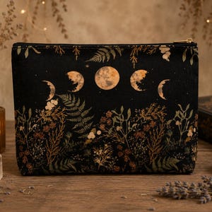 Op de afbeelding: Een zwarte rechthoekige etui met ritssluiting, voorzien van een goudkleurige print van de maanfasen aan de bovenzijde en een gedetailleerd botanisch patroon van varens en diverse wilde planten op het onderste deel van de stof.