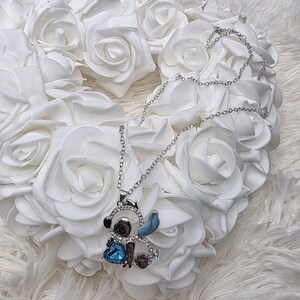 May include: Silver-toned necklace featuring a cartoon character pendant. The pendant has a blue heart-shaped gem and is embellished with clear crystals. The necklace is displayed on a bed of white roses.