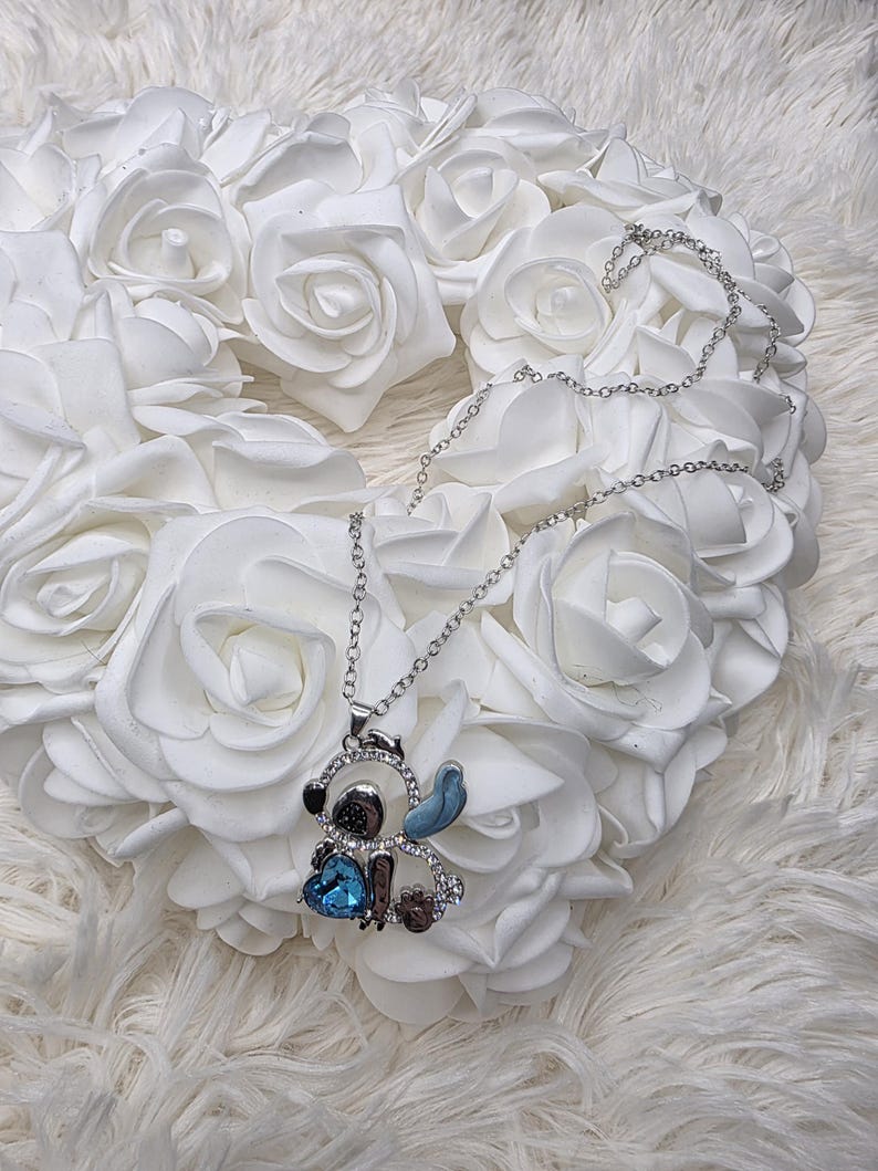 May include: Silver-toned necklace featuring a cartoon character pendant. The pendant has a blue heart-shaped gem and is embellished with clear crystals. The necklace is displayed on a bed of white roses.