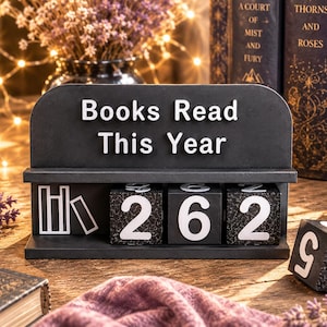 Könnte beinhalten: Ein schwarzer Holzaufsteller mit dem weißen Text Books Read This Year, der ein Regal für drei nummerierte Blöcke und einen separaten losen Block enthält. Die Blöcke zeigen weiße Zahlen auf schwarzem, strukturiertem Grund und ein Buchsymbol.