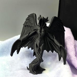 May include: A black figurine of a cloaked figure with large, detailed wings. The figure has a hood, horns, and a flowing robe. The statue is a gothic or fantasy-themed item, likely a decorative piece or collectible.