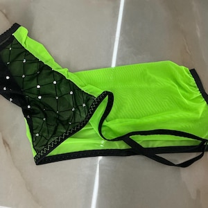Könnte beinhalten: Ein neon-grünes Netz-Kleidungsstück mit einem schwarzen Netzeinsatz auf einer Seite, der ein rautenförmiges Gittermuster und kleine eingebettete Strasssteine aufweist. Das Teil ist mit schwarzer Einfassung und dünnen schwarzen Riemen versehen.
