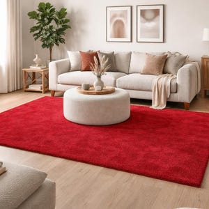 Puede incluir: Una gran alfombra rectangular roja domina el primer plano de un salón. También hay un sofá de color claro con cojines y una otomana redonda. La habitación es luminosa, con luz natural.