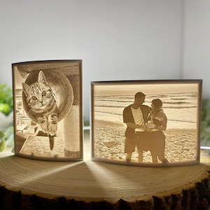 Könnte beinhalten: Zwei beleuchtete Fotoblöcke. Einer zeigt eine Katze, die nach unten schaut, der andere ein Paar am Strand. Die Blöcke sind rechteckig mit gewölbten Oberteilen und sitzen auf einer Holzscheibe. Die Bilder sind in Sepiatönen gehalten.