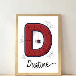 May include: Framed print featuring a large red letter "D" with a spiderweb design and a small spider hanging from a web. The name "Dustine" is written in a cursive font below the letter. The frame is light brown.