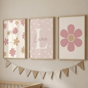 May include: Three framed floral art prints in a nursery setting. The prints feature pink and tan flowers, the letter "L" with the name "Luna", and a large pink flower. A garland of triangular flags hangs below the prints.