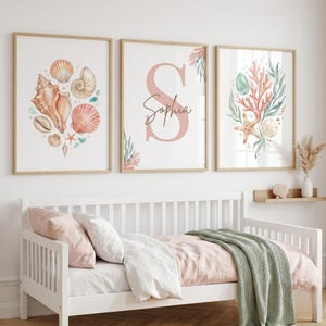 May include: Three framed wall art prints featuring watercolor illustrations of seashells, coral, and seaweed. The center print displays a large pink letter S with the name Sophia written in cursive script. The color palette includes soft pinks and greens.