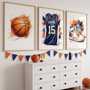 Può includere: Un set di tre stampe da parete incorniciate a tema basket. La prima stampa mostra un pallone da basket arancione, la seconda una maglia blu navy con il nome NOAH e il numero 15, la terza un paio di scarpe da ginnastica alte bianche e arancioni.