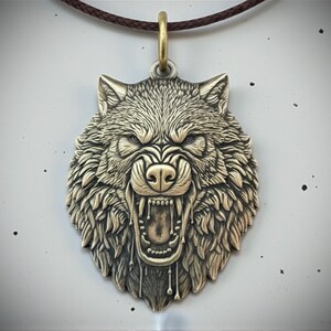 May include: A bronze-toned pendant depicting a snarling wolf's head. The pendant hangs from a brown cord with a gold-coloured clasp. The wolf's open mouth shows sharp teeth, and the fur is intricately detailed.